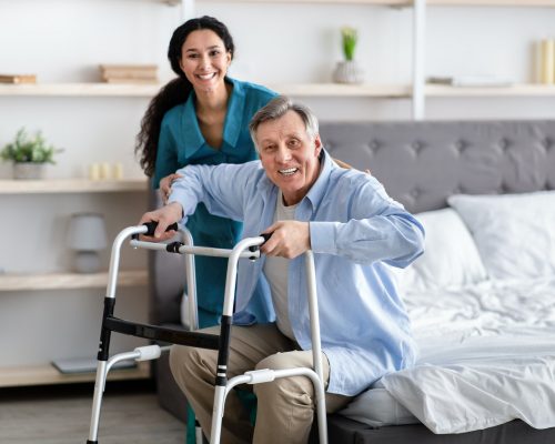 female-nurse-helping-elderly-male-with-walking-frame-stand-up-from-bed-at-home-professional-care